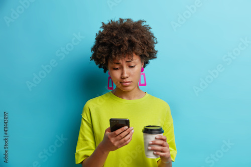 Displeased young pretty African American woman looks sadly at smartphone reads bad news in internet drinks takeaway coffee wears casual green t shirt and earrings isolated over blue background.