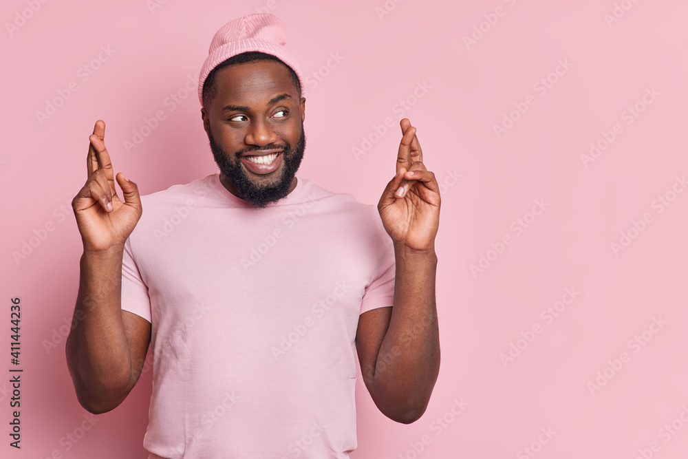 Wishing good luck. Positive dark skinned man with thick beard keeps fingers crossed dressed in casual clothes being in good mood isolated over rosy background copy space area. Wish in better Wishing good luck. Positive dark skinned man with thick beard keeps fingers crossed dressed in casual clothes being in good mood isolated over rosy background copy space area. Wish in better