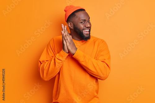 Fotografija Positive dark skinned man rubs palms and smiles gladfully has intention to do something wears hat and sweater isolated over vivid orange background