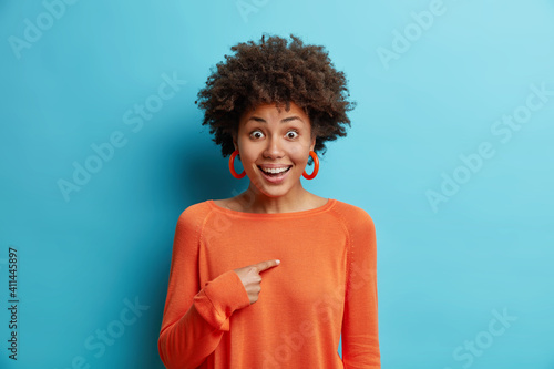 Cheerful surprised young pretty African American woman points at herself asks who me smiles broadly didnt expect being chosen dressed in long sleeved orange jumper isolated over blue background photo