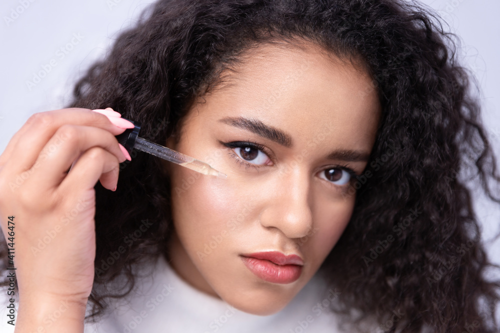 Obraz premium Close up mixed race curly young female applying skincare facial moisturizing nourishing serum or natural oil on skin around eye by pipette looking at camera, anti-wrinkle treatment.