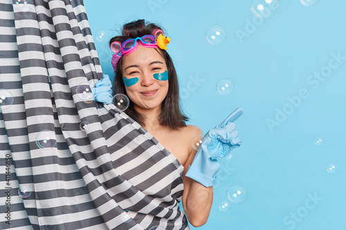 Enjoy simple things. Delighted brunette Asian woman applies collagen pads under eyes hides naked body behind shower curtain indicates aside on blue background with bubbles has healthy soft skin