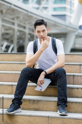 Front view portrait of a handsome young adult Asian man in casual white clothes and backpack sitting on the stair while holding a folding paper map and looking at the camera with a blurred background
