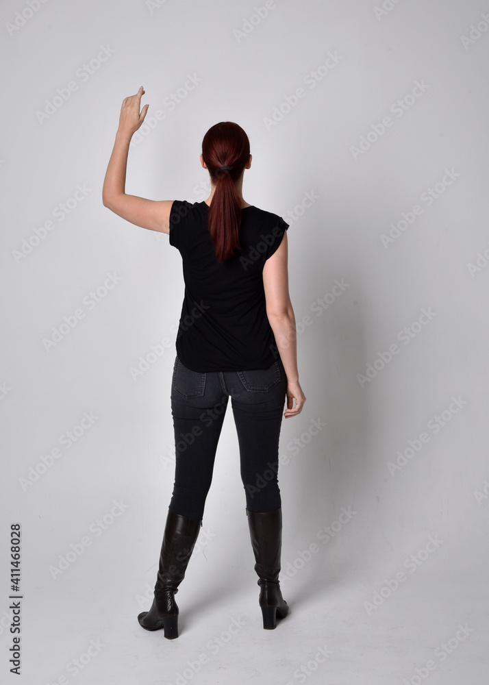 Full length portrait of woman with red hair in a ponytail, wearing black t-shirt and denim pants. Standing pose, hands reaching out with back to the camera the camera, against a studio background. Full length portrait of woman with red hair in a ponytail, wearing black t-shirt and denim pants. Standing pose, hands reaching out with back to the camera the camera, against a studio background.
