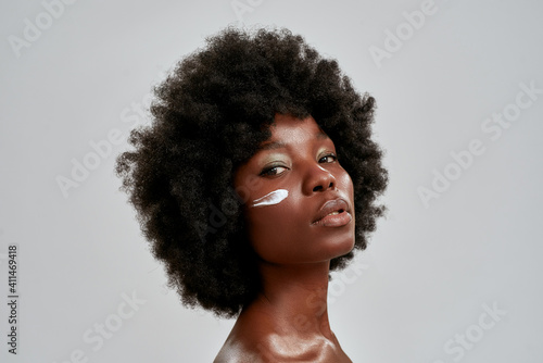 Beauty portrait of young naked african american woman with afro hair looking at camera while posing with cream applied on her face isolated over gray background
