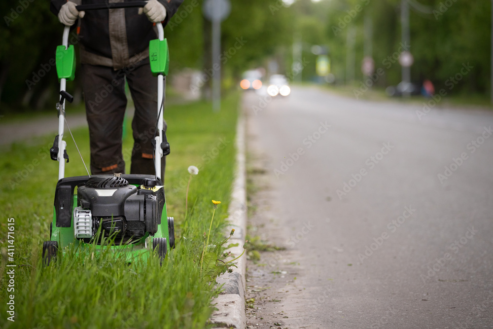 A worker in protective clothing with a gasoline lawn mower on wheels, mows the grass along the road. The powerful mower mows flowers and other plants. Territory care.