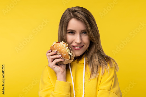 Photo of attractive lady poses in a cute way with burger and smile. Wears casual yellow hoody, isolated yellow color background