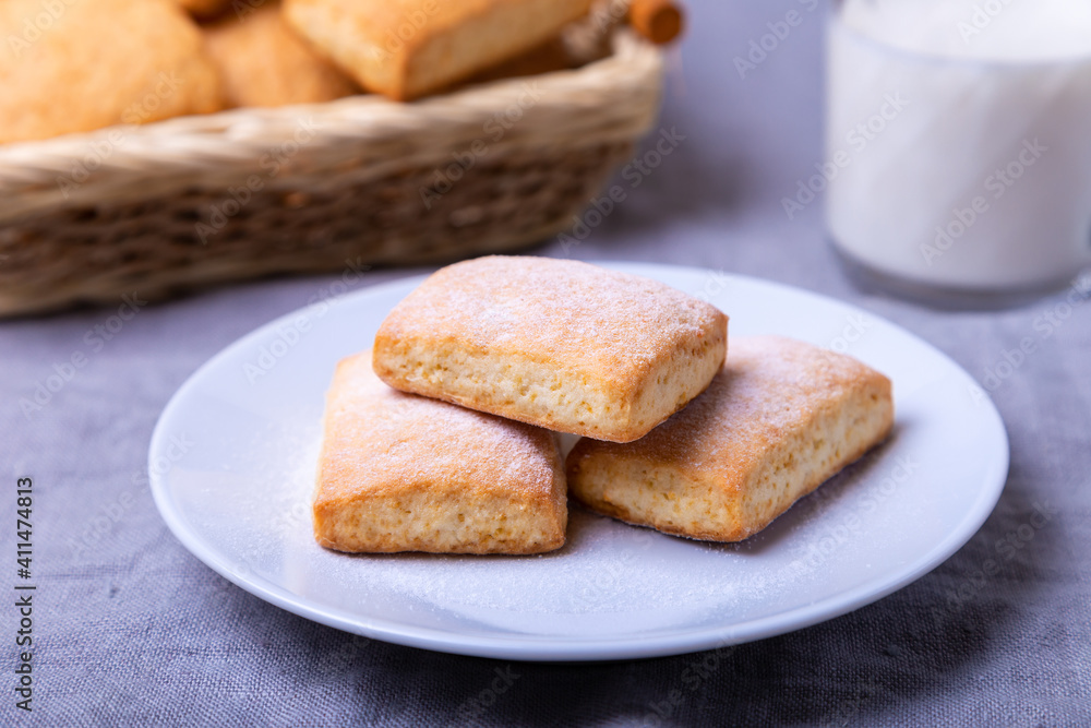 Cookies with powdered sugar on a white plate. In the background a basket of cookies and a cup of milk. Close-up. Cookies with powdered sugar on a white plate. In the background a basket of cookies and a cup of milk. Close-up.