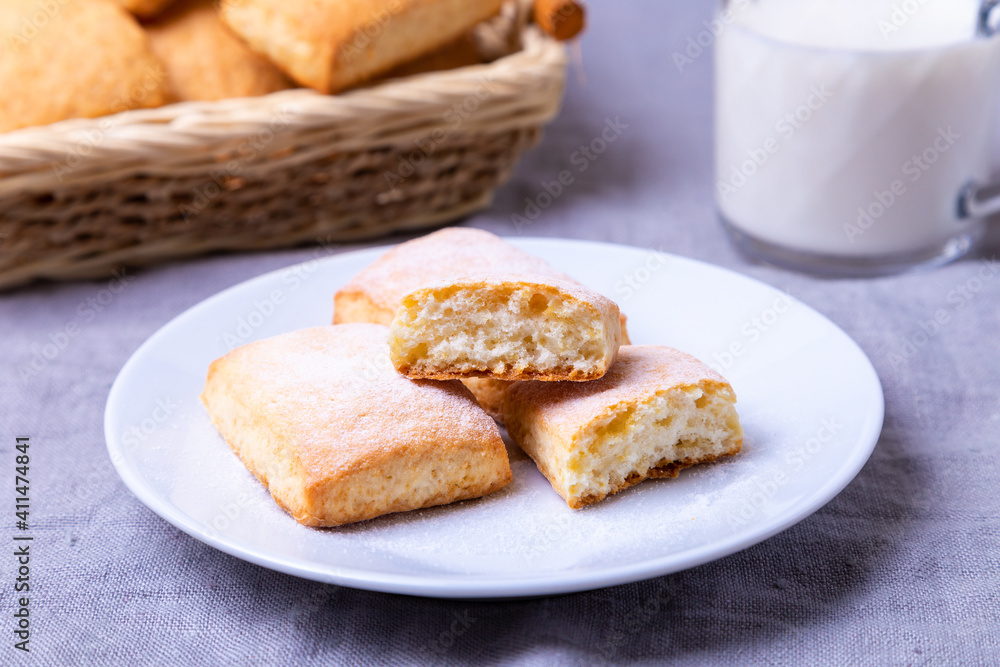 Cookies with powdered sugar on a white plate. In the background a basket of cookies and a cup of milk. Close-up. Cookies with powdered sugar on a white plate. In the background a basket of cookies and a cup of milk. Close-up.