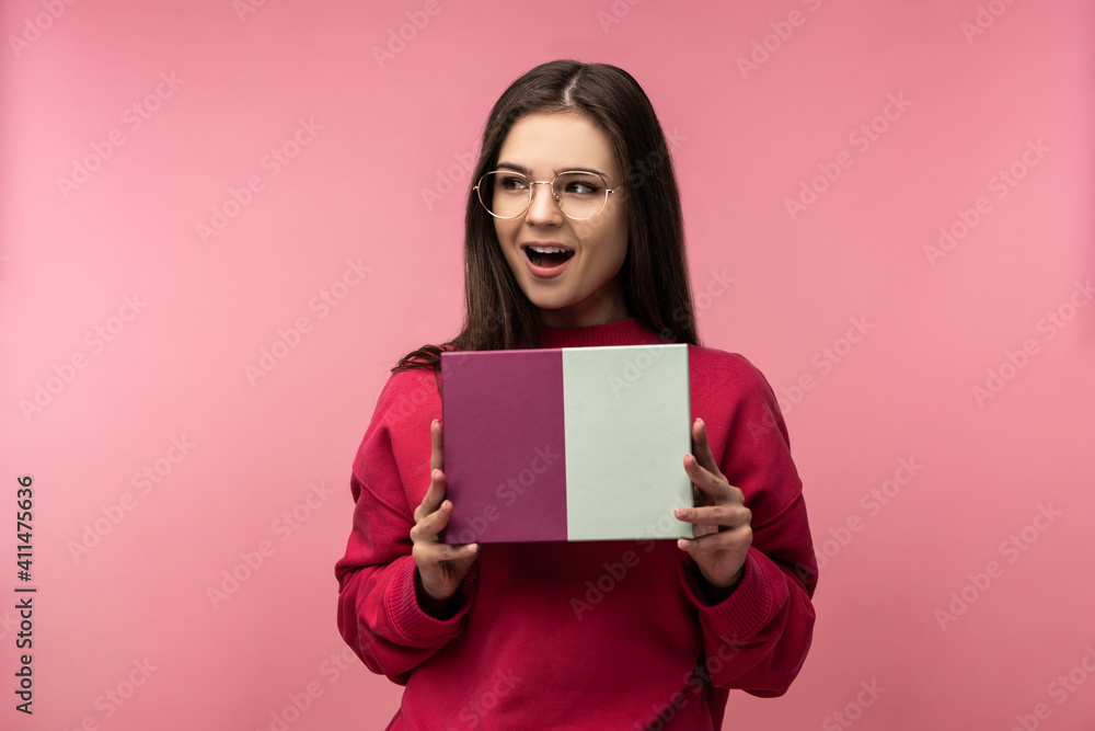 Photo of attractive lady in glasses holds box with present, waits and looks forward to open. Wears casual pink sweater white pants isolated pink color background Photo of attractive lady in glasses holds box with present, waits and looks forward to open. Wears casual pink sweater white pants isolated pink color background