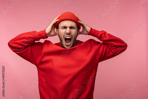 Photo of attractive man with beard in glasses and red clothing. Male screams and yells holds his head, isolated over pink background © Ivan