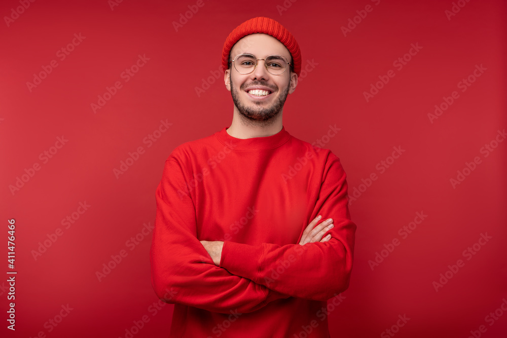 Photo of attractive man with beard in glasses and red clothing. Male crossed hands and smiles, isolated over red background Photo of attractive man with beard in glasses and red clothing. Male crossed hands and smiles, isolated over red background