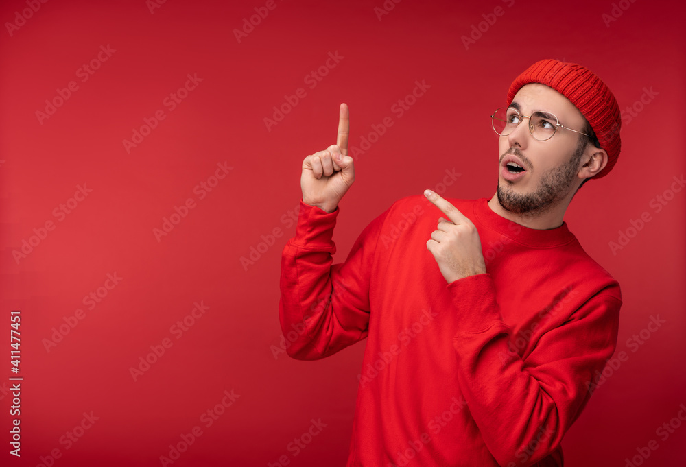 Naklejka premium Photo of attractive man with beard in glasses and red clothing. Shocked and puzzled male points up, isolated over red background