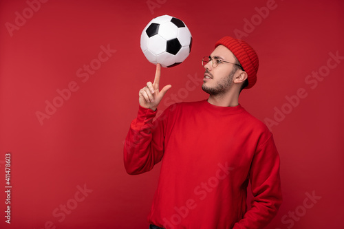 Photo of attractive man with beard in glasses and red clothing. Happy man holds the ball on the index finger, isolated over red background