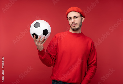Photo of attractive man with beard in glasses and red clothing. Happy man holds ball, isolated over red background