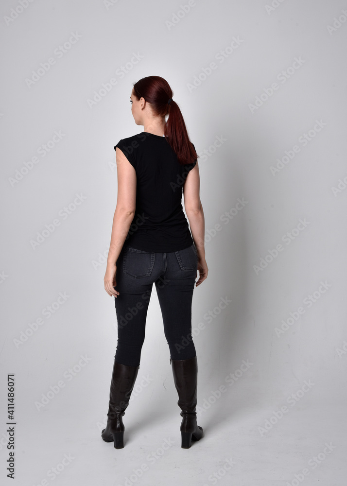 Simple full length portrait of woman with red hair in a ponytail, wearing casual black tshirt and jeans. Standing pose with back to the camera the camera, against a studio background. Simple full length portrait of woman with red hair in a ponytail, wearing casual black tshirt and jeans. Standing pose with back to the camera the camera, against a studio background.