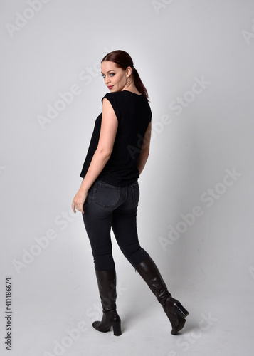 Simple full length portrait of woman with red hair in a ponytail, wearing casual black tshirt and jeans. Standing pose with back to the camera the camera, against a  studio background. © faestock