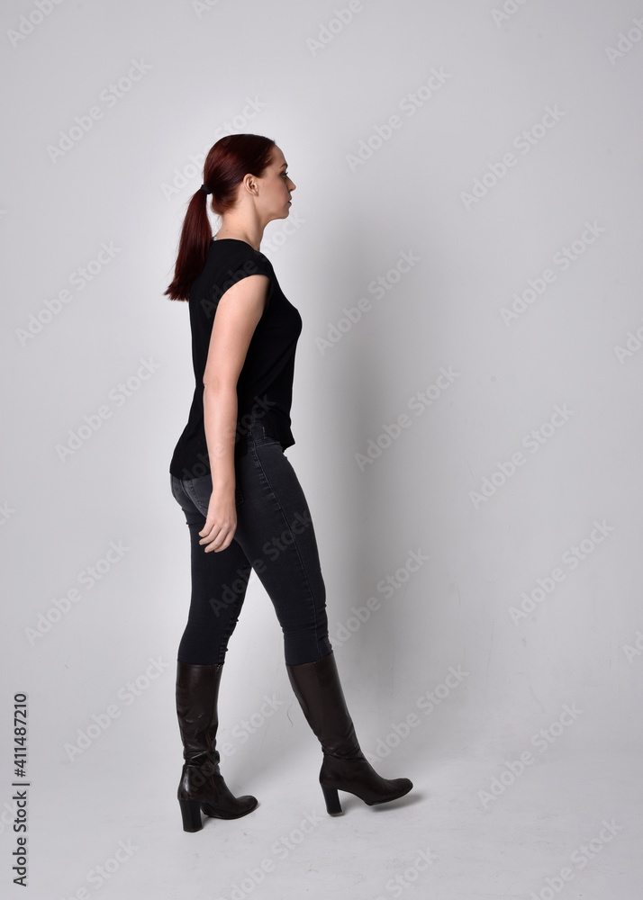 Simple full length portrait of woman with red hair in a ponytail, wearing casual black tshirt and jeans. Standing pose with back to the camera the camera, against a studio background. Simple full length portrait of woman with red hair in a ponytail, wearing casual black tshirt and jeans. Standing pose with back to the camera the camera, against a studio background.