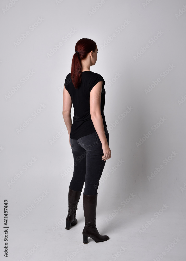 Simple full length portrait of woman with red hair in a ponytail, wearing casual black tshirt and jeans. Standing pose with back to the camera the camera, against a studio background. Simple full length portrait of woman with red hair in a ponytail, wearing casual black tshirt and jeans. Standing pose with back to the camera the camera, against a studio background.