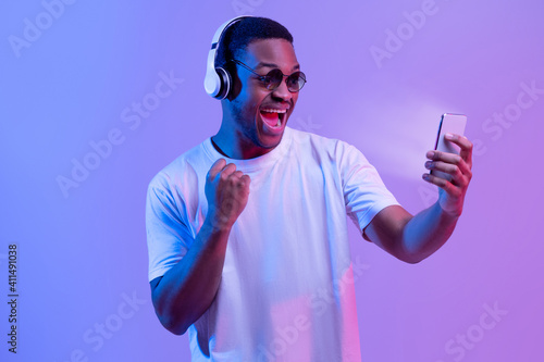 Online Gaming. Excited Black Guy In Headphones Celebrating Success With Smartphone