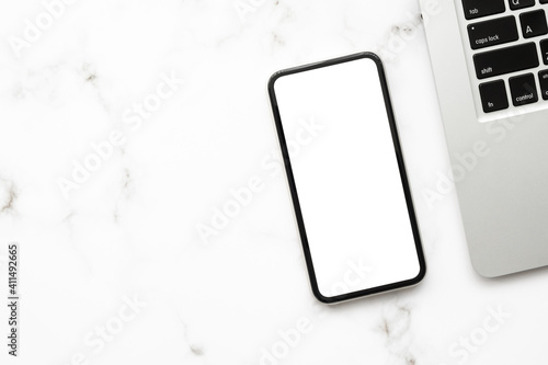 Smartphone with blank screen is on top of white marble office desk table with laptop computer. Top view with copy space  flat lay.