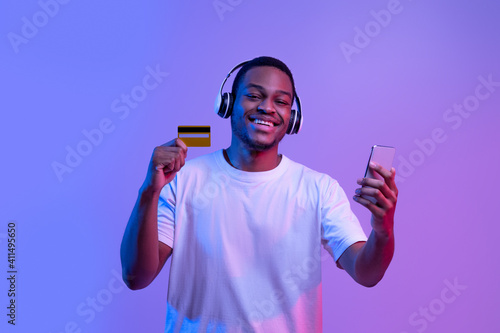 Streaming Music Subscription. Happy black guy in headphones holding smartphone and card photo