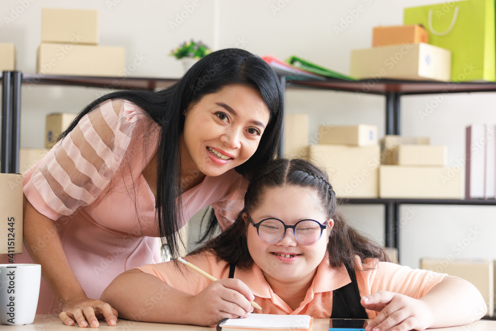 Middle-aged mother with an Asian autistic daughter is looking at the camera and smiling inside a home converted into a small business office online. New business in the era of the COVID-19 epidemic.