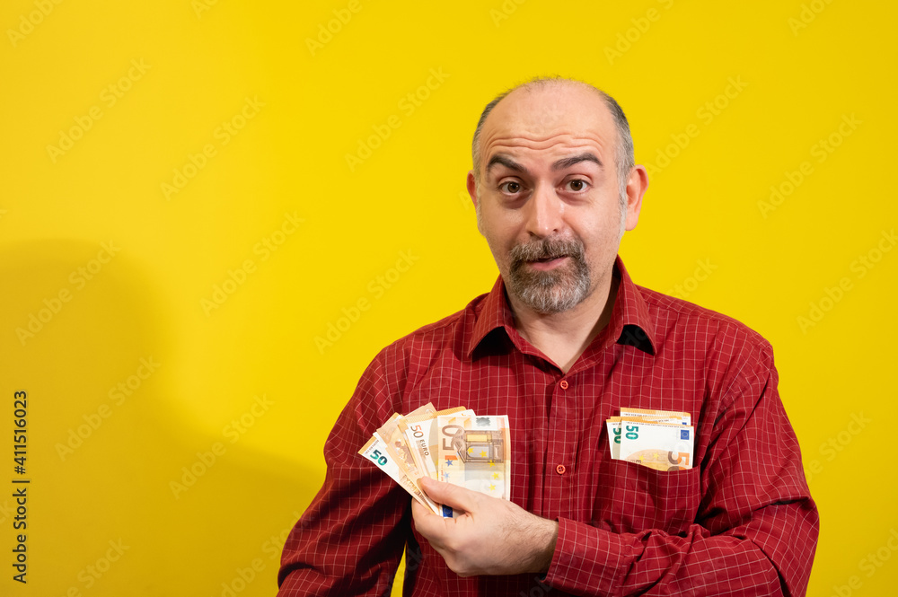 Middle-aged Caucasian man holding a bunch of 50 euro bills in his hand and has more in his shirt pocket. He is happily surprised. Concept of winning, luck and wealth. Yellow background.