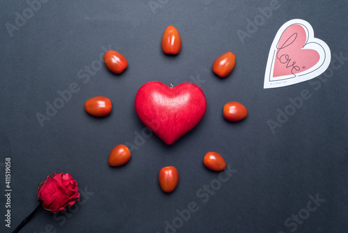 Top view on a red love heart, surrounded by small tomatoes, looking like a shining sun or a flower, with a paper heart and a red rose next to it, black background. Concept of love and valentine's day