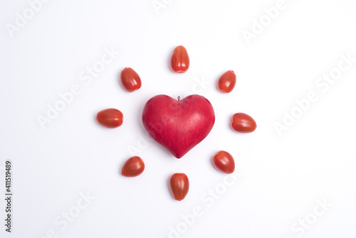 Top view on a red love heart, surrounded by small tomatoes and looking like a shining sun or a flower, white background. Concept of love and valentine's day
