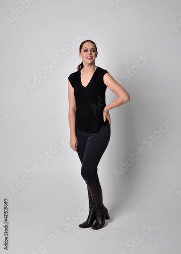 Simple full length portrait of woman with red hair in a ponytail  wearing casual black tshirt and jeans. Standing pose facing front on  against a  studio background.