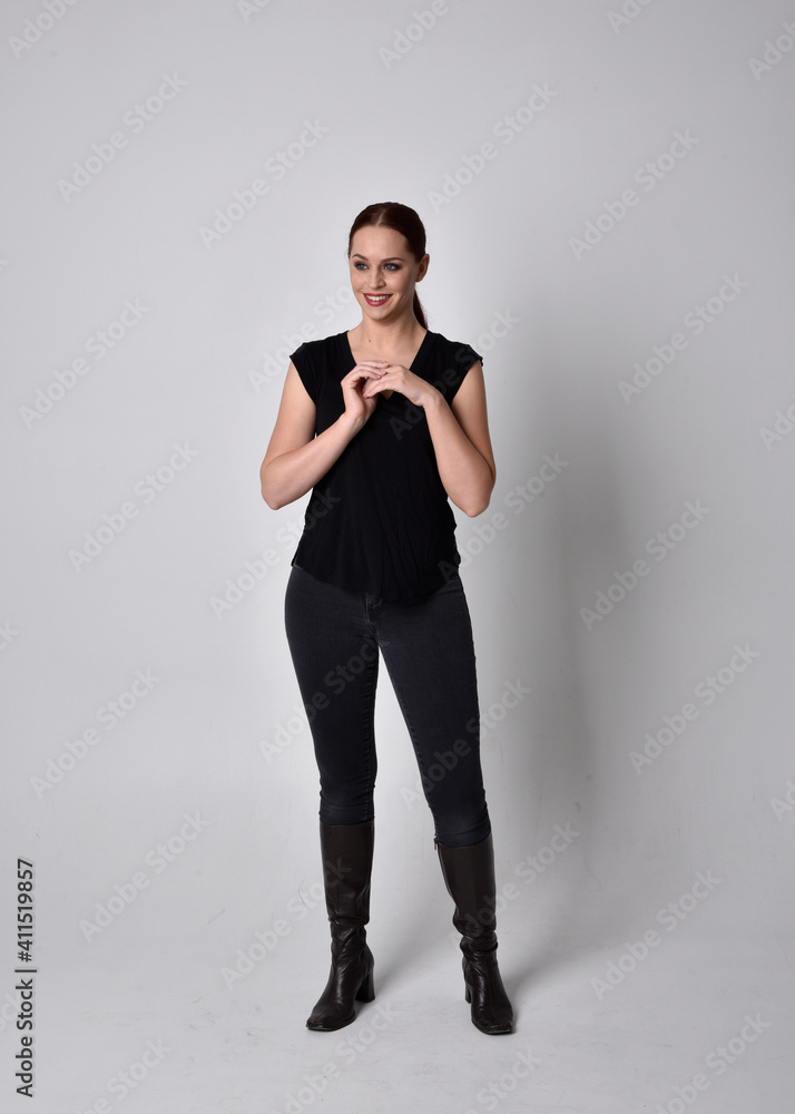 Simple full length portrait of woman with red hair in a ponytail, wearing casual black tshirt and jeans. Standing pose facing front on, against a studio background. Simple full length portrait of woman with red hair in a ponytail, wearing casual black tshirt and jeans. Standing pose facing front on, against a studio background.