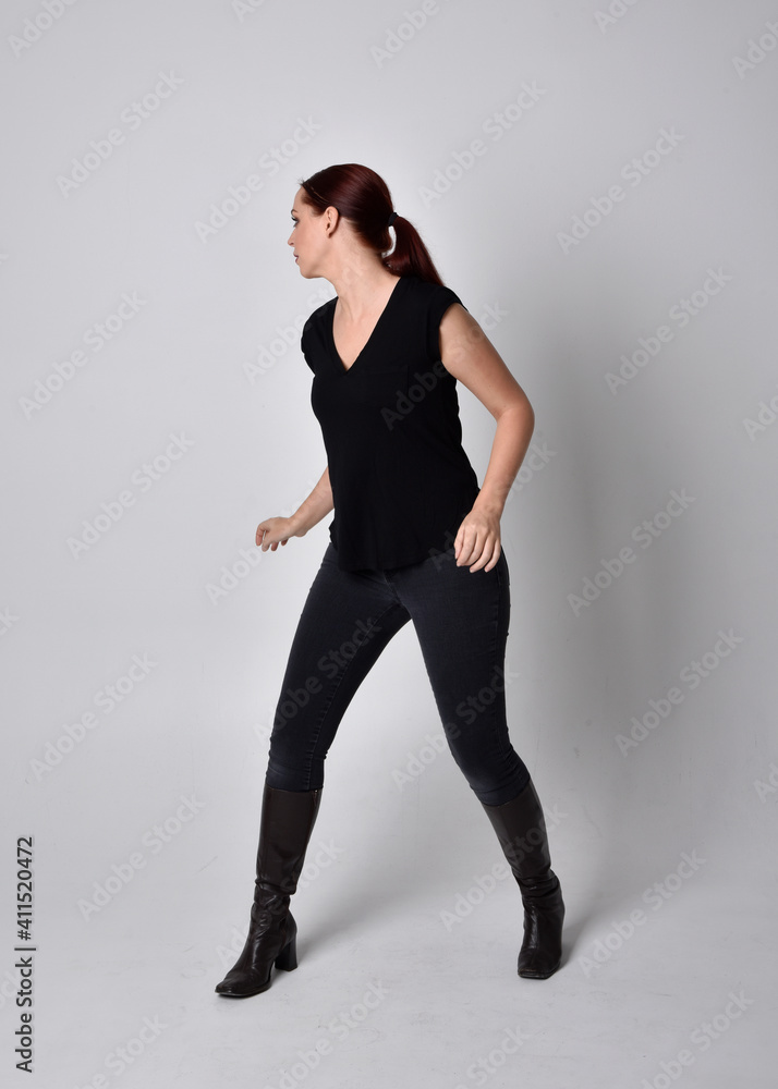 Obraz premium Simple full length portrait of woman with red hair in a ponytail, wearing casual black tshirt and jeans. Standing pose facing front on, against a studio background.