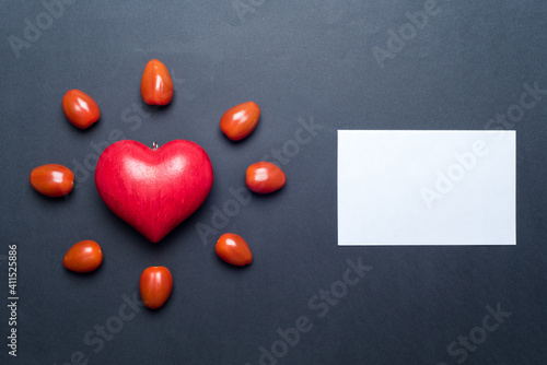 Red love heart, surrounded by small tomatoes, looking like a shining sun or a flower, with a blank gift card next to it, black background. Concept of love and valentine's day