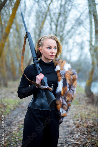 caucasian woman in black suit, corset and fur on the shoulder with shotgun on the shoulder in the autumn forest