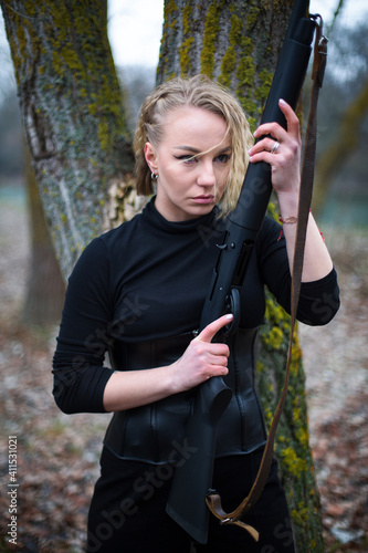 blonde woman in black suit and corset with shotgun stays in front of tree trunk in the autumn forest