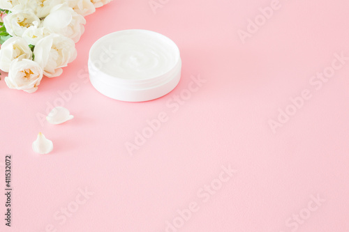 Opened white jar of natural herbal cream for women on light pink table. Pastel color. Beautiful roses. Fresh flowers. Care about clean and soft face, hands, legs and body skin. Empty place for text.