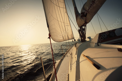 White sloop rigged yacht sailing in an open sea at sunset. Clear sky. A view from the deck to the bow, mast, sails. Transportation, travel, cruise, sport, recreation, leisure activity, racing, regatta photo