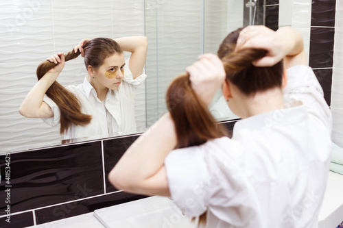 Young woman with hydrogel patches under eyes makes ponytail looking into the mirror in bathroom