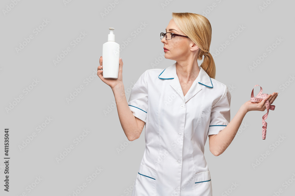 Portrait of beauty specialist standing against grey background and holding in her hand bottle of body lotion and measure tape Portrait of beauty specialist standing against grey background and holding in her hand bottle of body lotion and measure tape