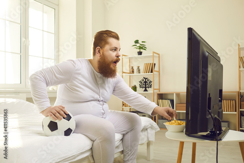 Funny emotional red haired bearded overweight man in home pajamas watching football match on television at home and eating popcorn. Funny male look concepts