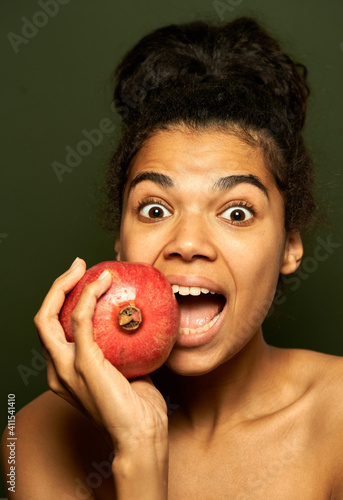 More fruits more life. Portrait of young mixed race woman looking surprised, holding pomegranate fruit near her mouth, posing isolated over green background. Healthy eating, vitamins concept