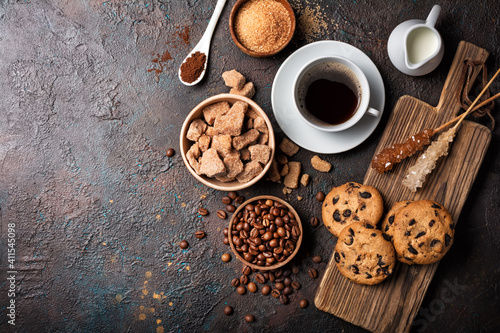 Cup of coffee with chocolate drop cookies, bowls of brown cane lump and granulated sugar, crystal sugar sticks, milk and coffee beans
