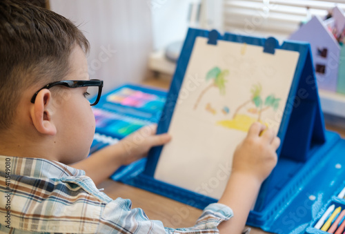 cute European boy with glasses draws on paper with pencils, sitting at the table. the process of creating a children's drawing