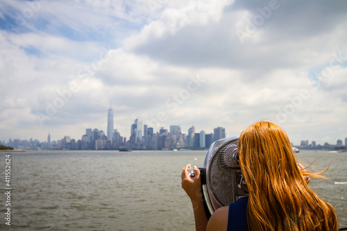 A young woman with red hair is looking at the cityscape of New York City through a binocular on Liberty Island