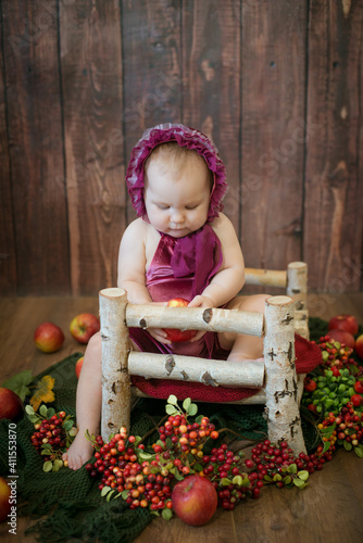 Little sweet girl up to one year old in a beautiful burgundy velvet suit with red juicy apples on a crib made of wood. Healthy lifestyle 