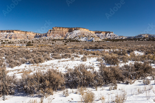 Snow covered mountains behind wide open desert vista covered in snow with brush on clear day in rural New Mexico