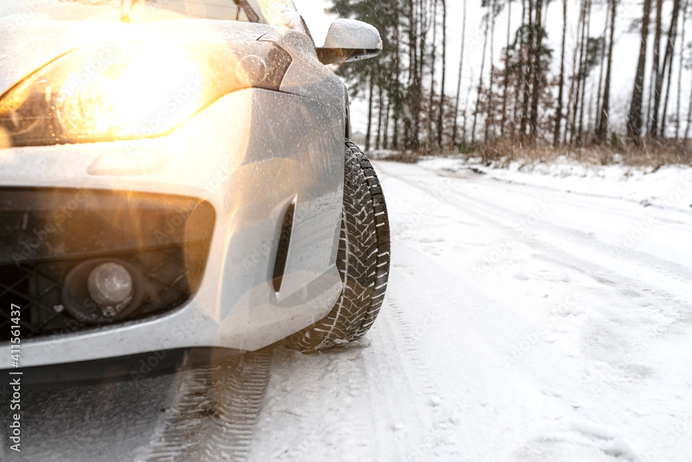 The dipped headlights on a silver car standing on a snowy road in the forest, visible new winter tire and falling snow.