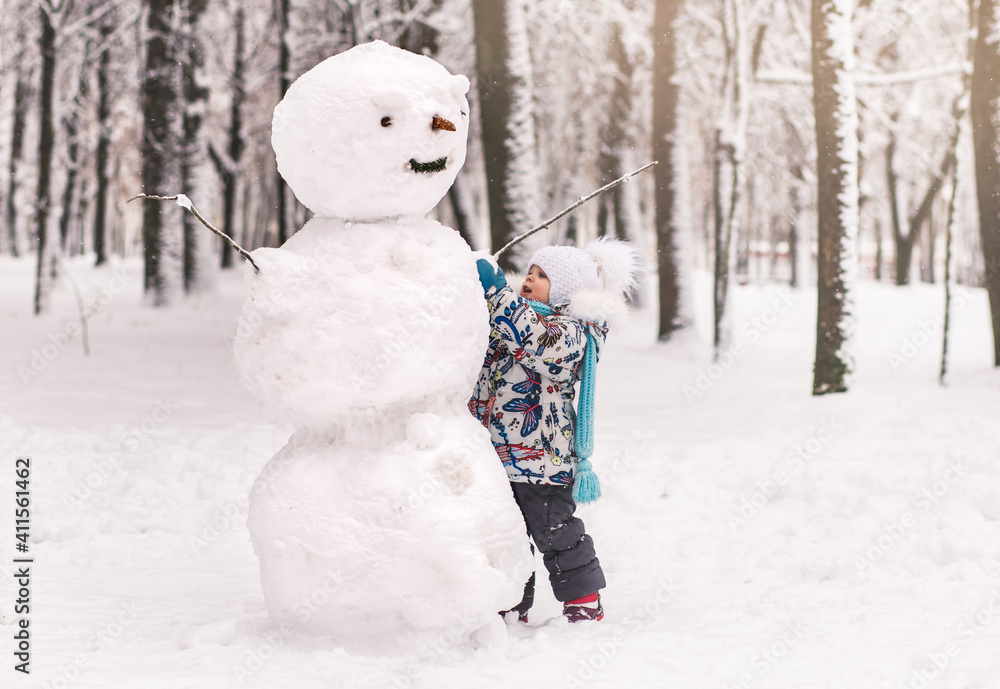 Little cute girl sculpts a big snowman in winter in the park