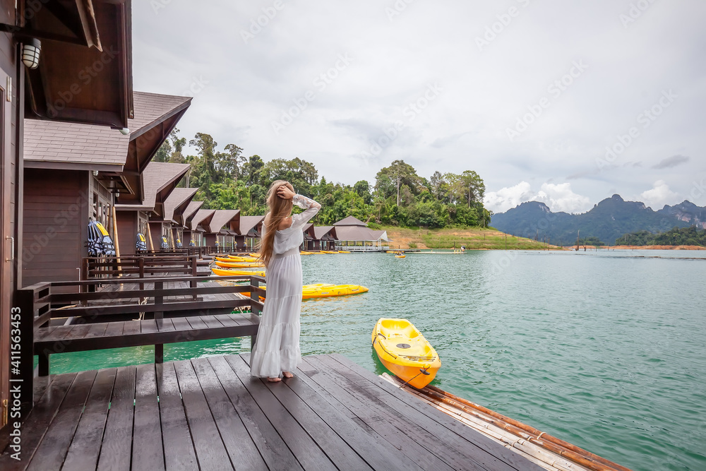 Young Travel Woman in White Dress Standing on Pier of Wooden House on Green Lake with Tropical Mountains. Female Tourist in Luxury Resort with Floating Raft Bungalows at Cheow Lan Lake, Thailand